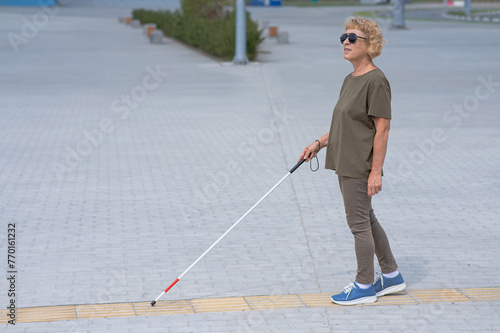 An elderly blind woman walks with a cane along a tactile tile. 
