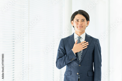 オフィスで胸に手を当てるスーツ姿の若い男性ビジネスマン（誠実・信頼・お任せください） 