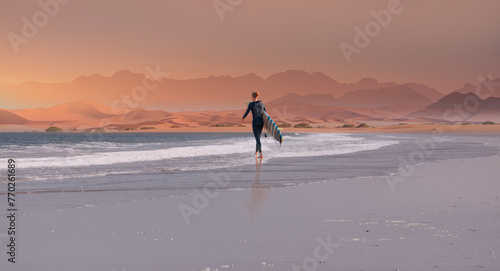 A male surfer walks on the beach with a surfboard in hand - Namib desert with Atlantic ocean meets near Skeleton coast - Namibia, South Africa