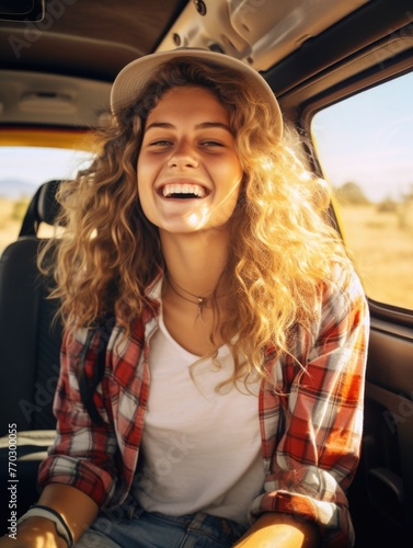 A woman with blonde hair and a red and white plaid shirt is smiling in a car. She is wearing a white shirt and a hat