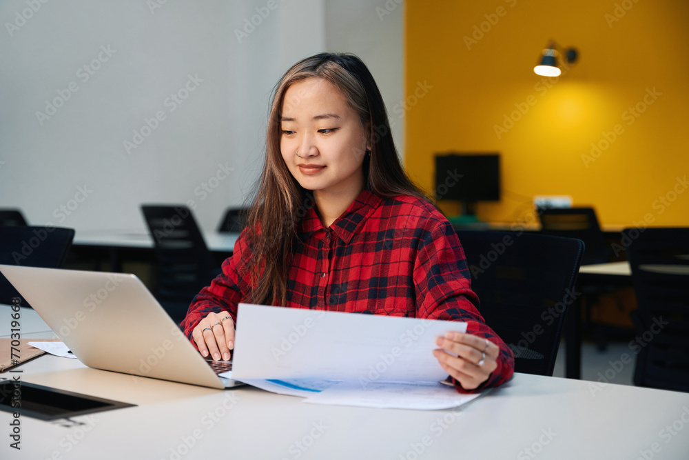 Young concentrated asian female IT employee with paper watching laptop at table Young concentrated asian female IT employee with paper watching laptop at table