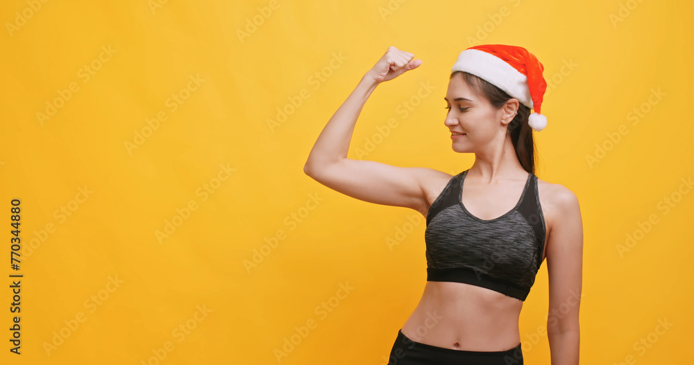 Young sportswoman wearing a Santa hat shows physical strength. Isolated on yellow background in studio. Young sportswoman wearing a Santa hat shows physical strength. Isolated on yellow background in studio.