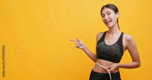 Young female in sportswear using a tape measure to measure her waist and pointing at something. Isolated on yellow background in studio.