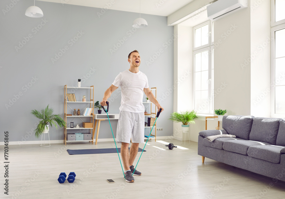 Young purposeful man at home doing sports strength training using rubber resistance band. Man in t-shirt and shorts shouting while stretching elastic band in living room. Home fitness training concept Young purposeful man at home doing sports strength training using rubber resistance band. Man in t-shirt and shorts shouting while stretching elastic band in living room. Home fitness training concept