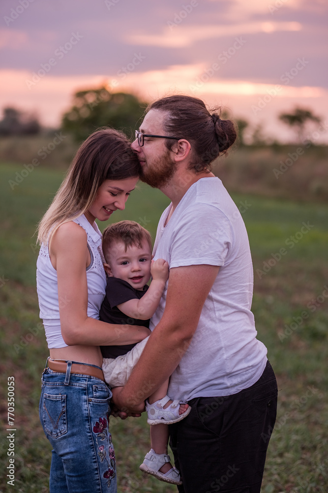 Fototapeta premium Diversity stylish father kisses mothers forehead while holding little son, concept of beautiful family connection against vibrant sunset. Family ties, togetherness. Close up portrait