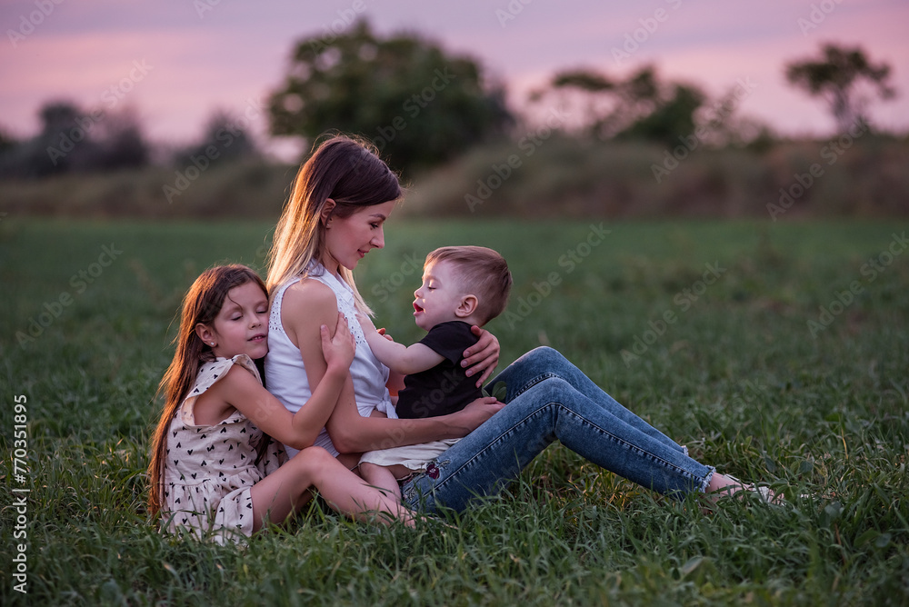 Mother embraces son and daughter, sitting on grassy field as twilight settles. Moment of tranquil affection, connection. Serene Family Time at Dusk. Woman hugging kids in green meadow. Mothers day Mother embraces son and daughter, sitting on grassy field as twilight settles. Moment of tranquil affection, connection. Serene Family Time at Dusk. Woman hugging kids in green meadow. Mothers day