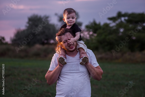 Diversity father with beard carrying son on shoulders shares tender moment in twilight lit field, embodying the essence of paternal love. Man and boy fooling around, playing. Child touches mans nose