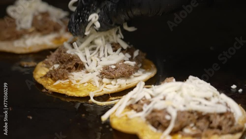 Shredded cheese added to birria tacos cooking on flat top grill, black gloved hand sprinkles grated quesadilla cheese to sizzling tacos, slow motion slider close up 4K photo