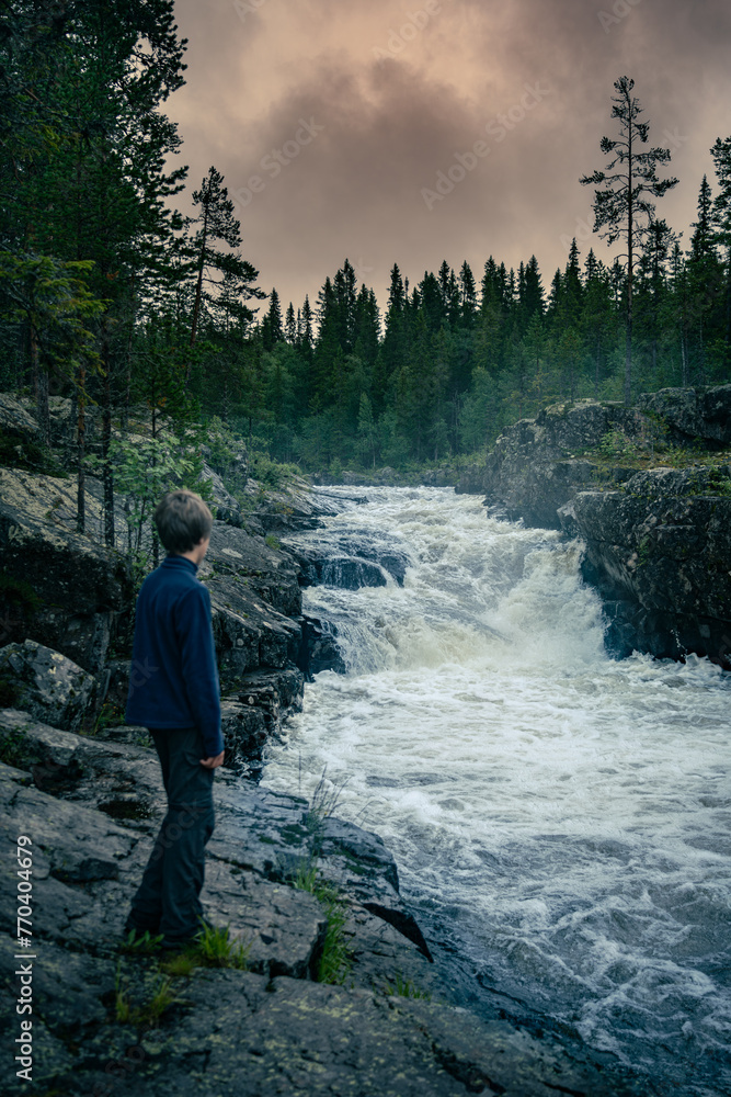 Boy Observing Thundering Sognstupet rapids in Idre Dalarna Storån Forest River at Dusk. Young explorer stands on rocky terrain, captivated by the powerful waterfall in Sweden under orange dramatic sky