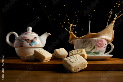 Taza de café con leche con tetera de flores y mariposas con fondo negro y salpicando unas pastas de té. photo
