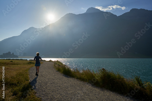 Early morning exercise at Lake Silvaplana. Landscape with water and mountain against the light. Woman jogging. Swiss, Sankt Moritz, Engadin.