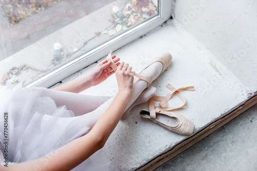 Overhead view captures a ballerina seated by a bright window, meticulously tying her pointe shoes in preparation for dance, surrounded by the urban scape outside.
