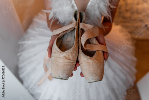 Young ballerina in white tutu, feathered hair tiara accessory stands thoughtfully, holding worn pointe shoes, evoking sense of dedication and the quiet before performance. White Swan
