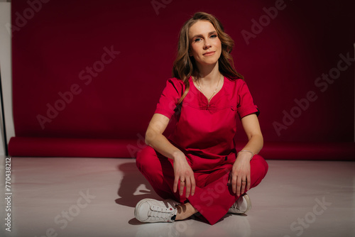 Young female nurse in a pink uniform sitting on the floor on a pink background