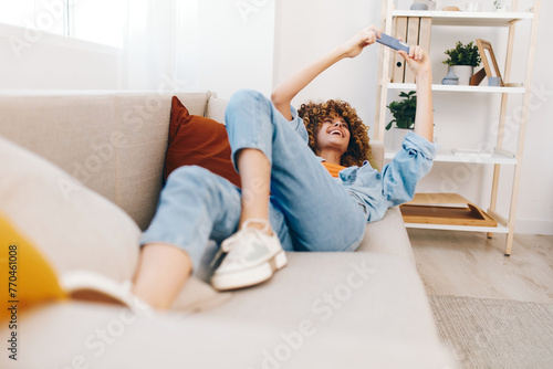 Smiling woman holding mobile phone, enjoying online games and reading messages on cozy couch in the living room.