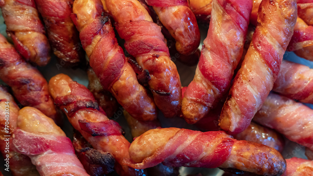 Sticky bacon-wrapped sausages spread on a plate to cool down, meat-based appetizer and popular party snack filling the frame, tasty protein-rich food that can be consumed in a carnivore or a keto diet Sticky bacon-wrapped sausages spread on a plate to cool down, meat-based appetizer and popular party snack filling the frame, tasty protein-rich food that can be consumed in a carnivore or a keto diet