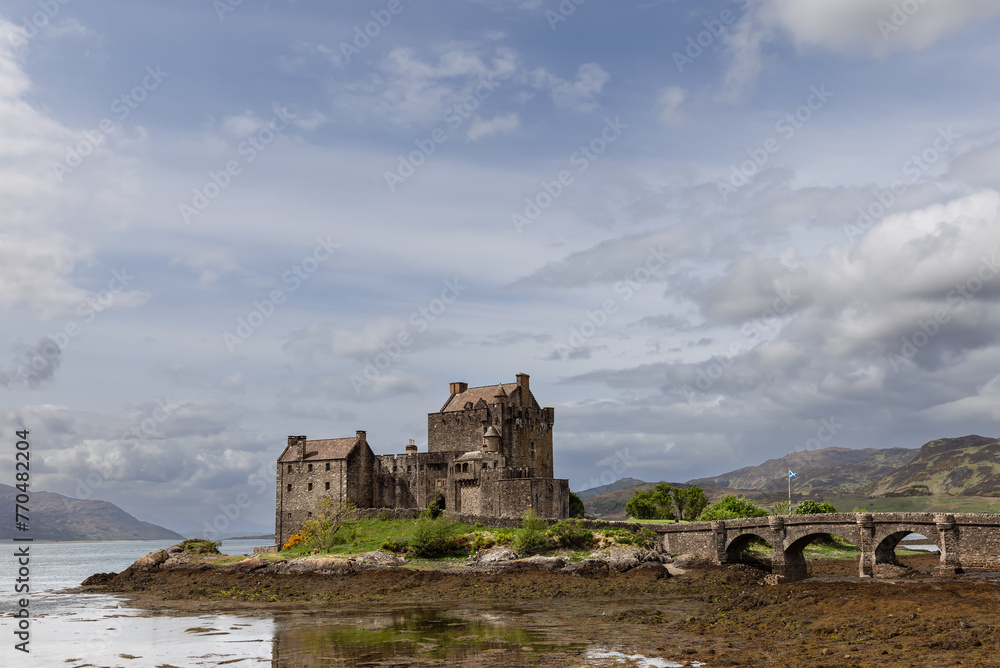 Eilean Donan Castle stands serene against a backdrop of rolling hills and dynamic skies, connected by an arched bridge reflecting in the tranquil lake waters