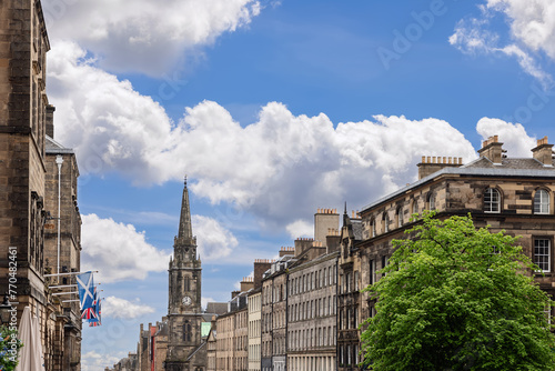 Edinburgh High Street comes to life in this picturesque scene, where the Scottish Saltire flutters above stone facades, and a steeple rises gracefully under the expansive, cloud-adorned sky