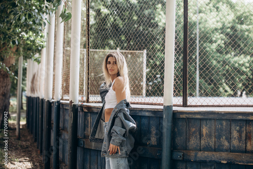 Beautiful blonde model, fashionable in jeans and a denim jacket, poses in the city against the background of a sports field grid