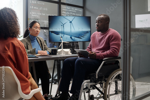 African American male architect with tablet sitting in wheelchair in front of female colleagues and making presentation of windmill energy