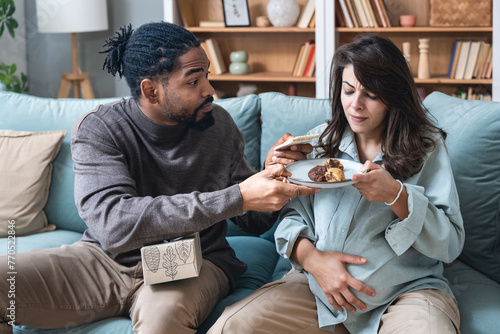 Pregnancy support and fun. Young couple man and pregnant woman sitting at home teasing with sweet food forbidden by doctor. Male and female support and love during pregnancy period enjoy together.