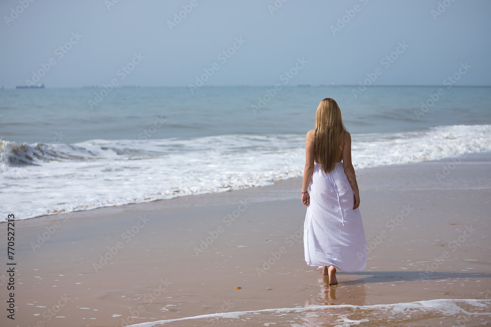 Young, beautiful, blonde woman in white dress, walking on a lonely beach, relaxed and calm, seen from the back. Concept peace, tranquility, solitude, finding oneself.