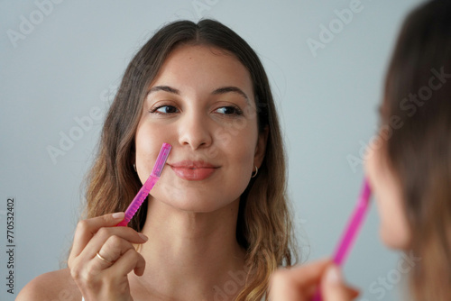 Dermaplaning. Close-up of beautiful girl shaving her face by razor at home. Pretty woman using razor on bathroom.