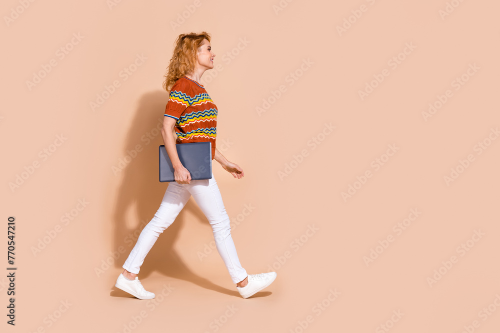 Photo portrait of lovely young lady hold netbook walking dressed stylish striped garment isolated on beige color background Photo portrait of lovely young lady hold netbook walking dressed stylish striped garment isolated on beige color background