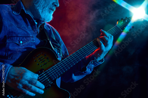 Senior bearded man playing guitar against dark background with dusk element. Concept, performance. Concept of music, instruments, concert, sound, equipment, festival photo