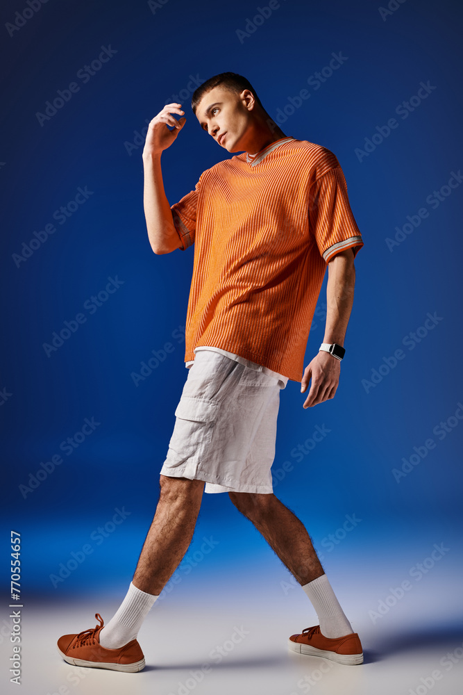 Full length image of handsome man in orange shirt and white shorts posing on blue backdrop