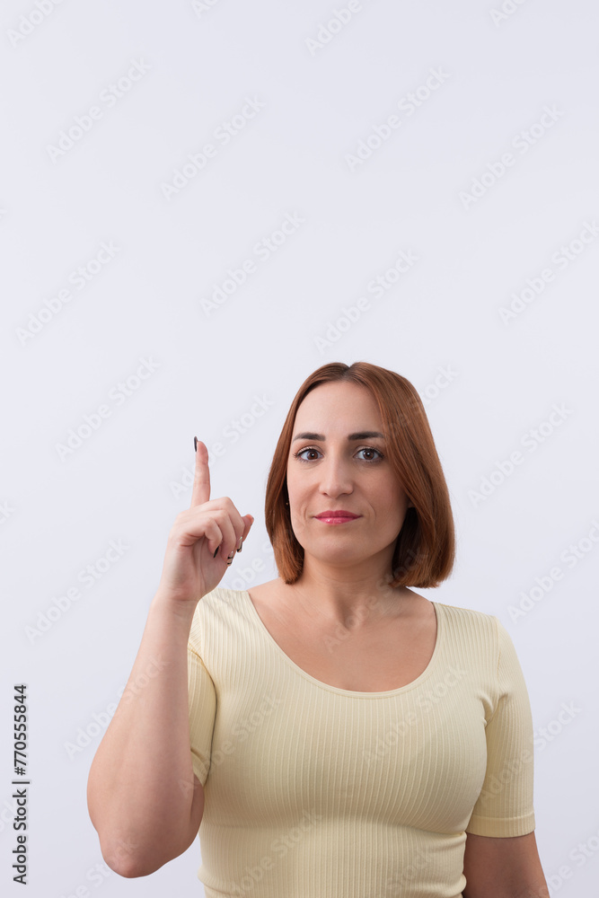 Beautiful young redheaded woman looking at camera smiling and pointing finger upward on light background. Copy space. Promotion and marketing concept Beautiful young redheaded woman looking at camera smiling and pointing finger upward on light background. Copy space. Promotion and marketing concept