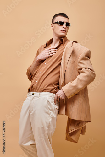 Fashion shot of man in beige jacket and shirt with sunglasses and hand near collar on beige backdrop