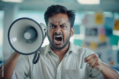 An angry Indian young man is talking on the loudspeaker of the phone he is holding in his hands. sitting in the office for recording an emotional voice, Generative AI © Story Sculptor