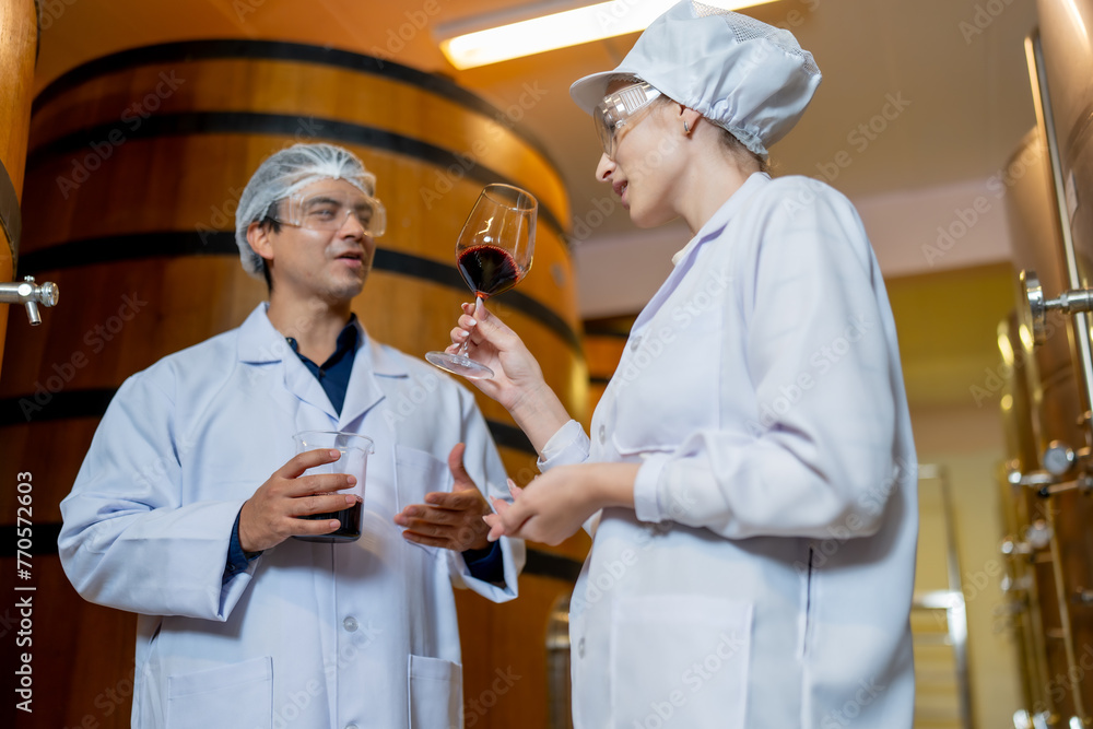 In a winery's aging room, a focused female wine expert evaluates the aroma of a wine sample while a male colleague observes.