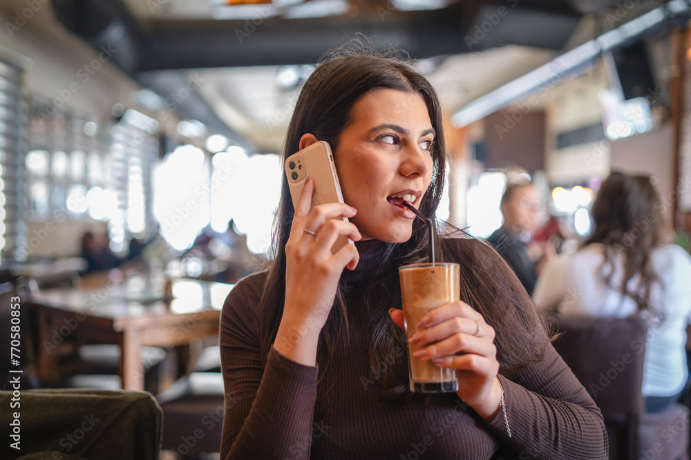A one young happy girl or woman is drinking cold coffee in cafe or restaurant while using her phone to send messages	
