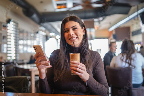 A one young happy girl or woman is drinking cold coffee in cafe or restaurant while using her phone to send messages  