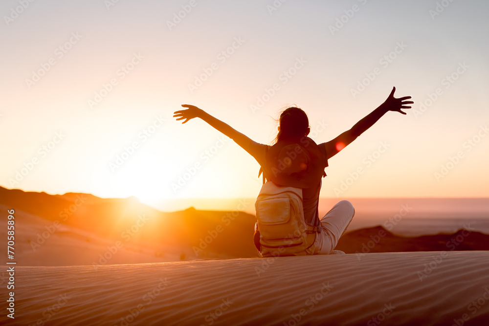 Happy girl woman with backpack is sitting with open arms against sunset desert