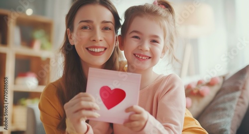 A woman and a little girl are holding a heart-shaped card. The woman is smiling and the girl is also smiling. The image conveys a warm and loving atmosphere