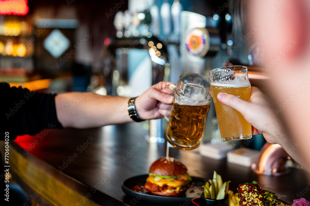 Two friends clinking beer glasses in a lively bar, with a burger and snacks in the foreground - obrazy, fototapety, plakaty 