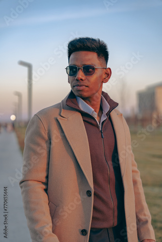 A man wearing a brown coat and sunglasses stands on a sidewalk
