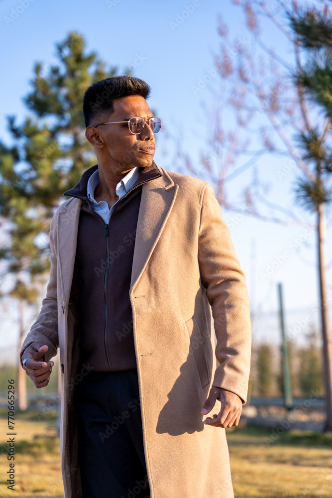A man wearing a brown coat and sunglasses is walking in a park. The man is wearing a white shirt and black pants A man wearing a brown coat and sunglasses is walking in a park. The man is wearing a white shirt and black pants