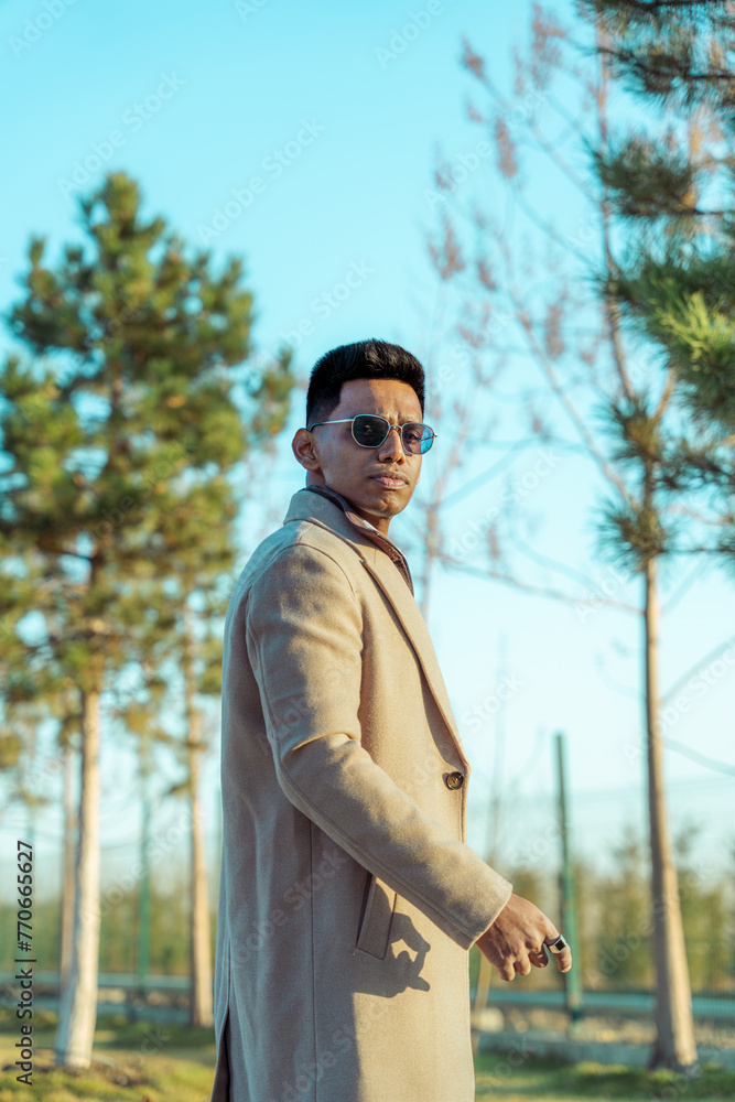 A man in a tan coat stands in front of a tree. He is wearing sunglasses and a tie A man in a tan coat stands in front of a tree. He is wearing sunglasses and a tie