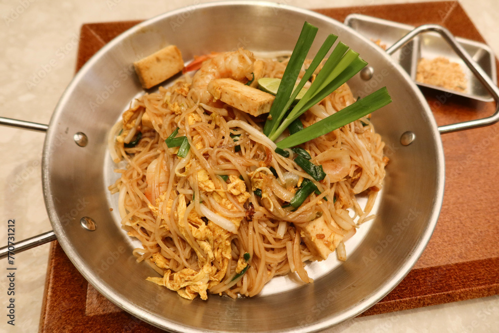 close up of prawn shrimp spring onion pad thai fried noodles in a steel pot dish and wooden tray. There is a bowl of salad and sauce on the side.