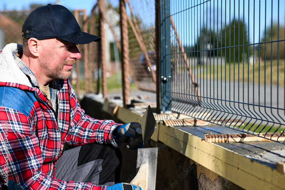 A man in overalls and gloves is repairing the fence in front of the family house. Close-up view and blurred background - obrazy, fototapety, plakaty 