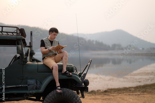 A young man contemplates sitting in his off-road vehicle by the lake. holding a notebook in the sunset