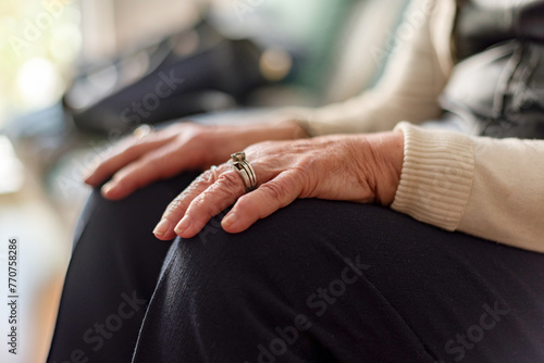 An elderly woman sits on a couch with her hands resting on her knees, wearing a ring on her left hand. Loneliness and old edge concept