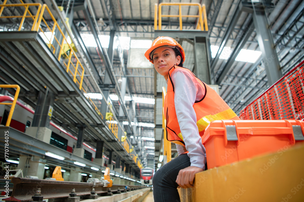 Portrait of female technician with toolbox, Sitting on a cement pedestal In front of the electric train tracks in the skytrain repair station.