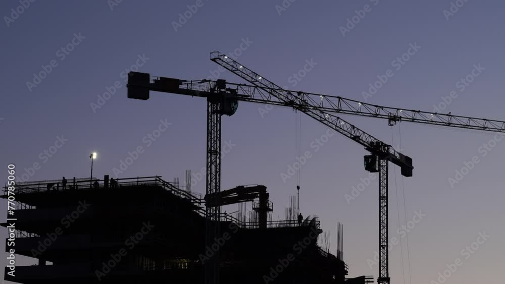 Illuminated tower cranes work at a construction site against a purple darkening sky. High quality 4k footage Illuminated tower cranes work at a construction site against a purple darkening sky. High quality 4k footage