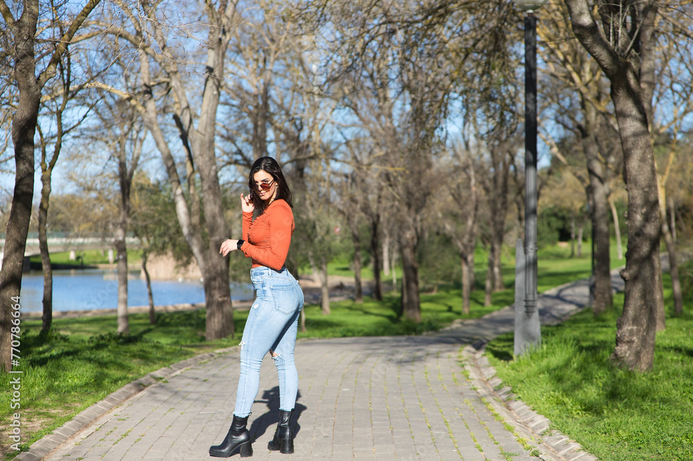 Young and beautiful brunette woman dressed in casual clothes and sunglasses, walks along a path between deciduous trees in autumn, in the background a lake. Young and beautiful brunette woman dressed in casual clothes and sunglasses, walks along a path between deciduous trees in autumn, in the background a lake.