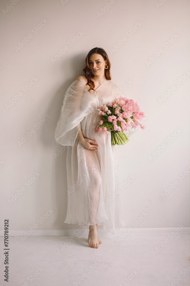 Portrait of pregnant woman in white transparent dress with bouquet of pink tulips posing near white wall in the studio. Copy space for text Portrait of pregnant woman in white transparent dress with bouquet of pink tulips posing near white wall in the studio. Copy space for text